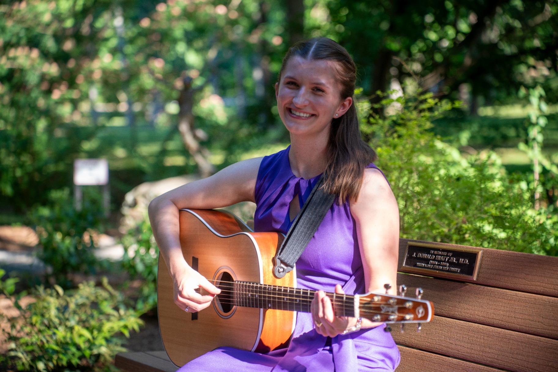 Music therapist in a purple dress smiling and playing acoustic guitar on a garden bench
