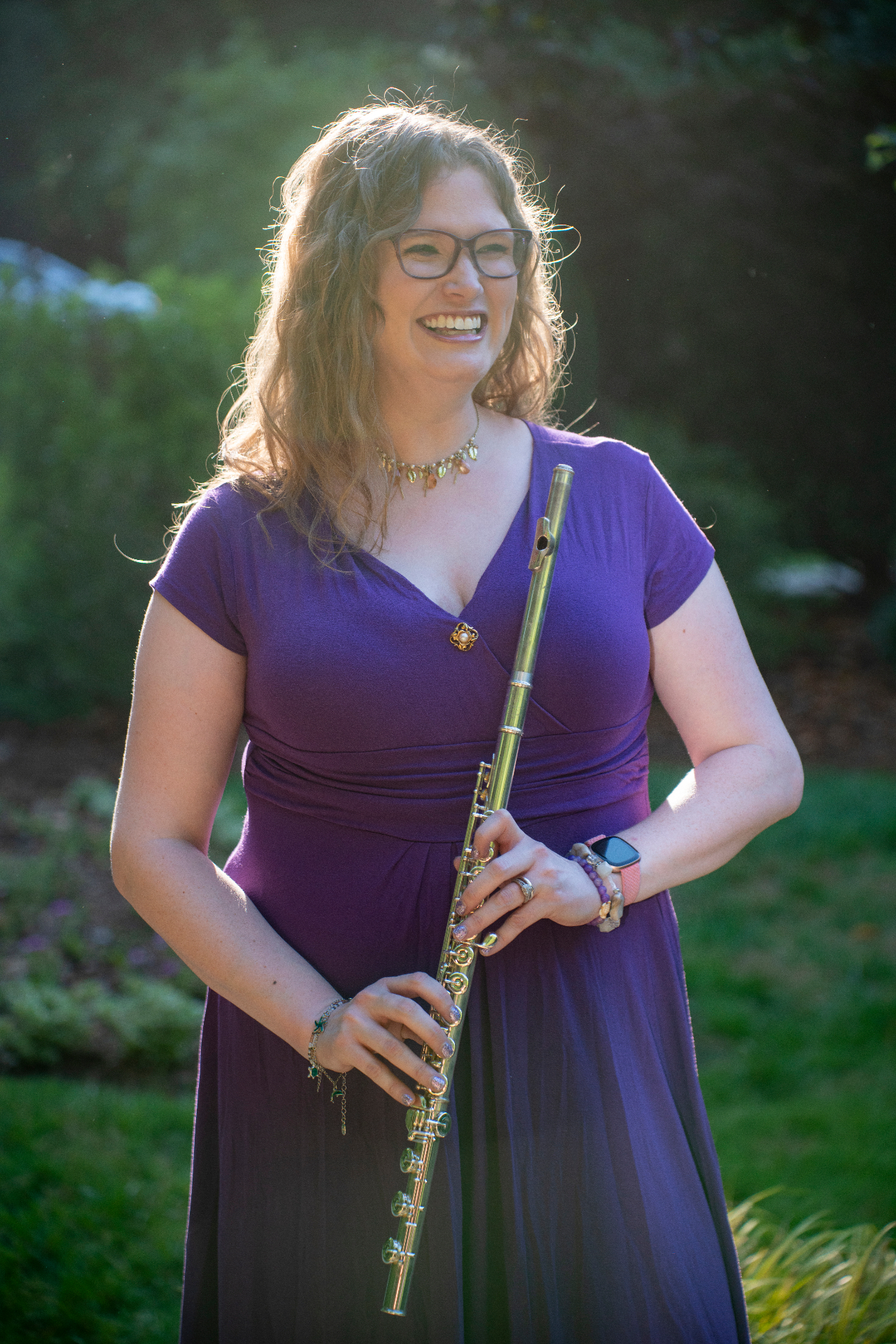Music therapist in a purple dress laughing while holding a silver flute in a sunlit garden