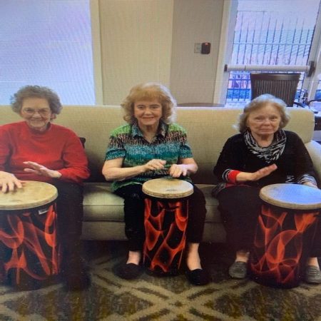 Three older adults seated together playing hand drums